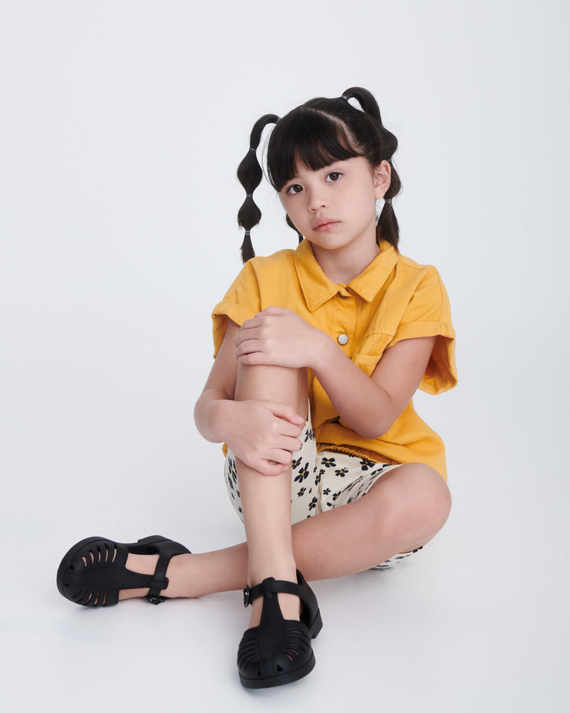 Child model in black and white shorts and yellow shirt wearing a pair of black Aranha 79 kids flats with buckle