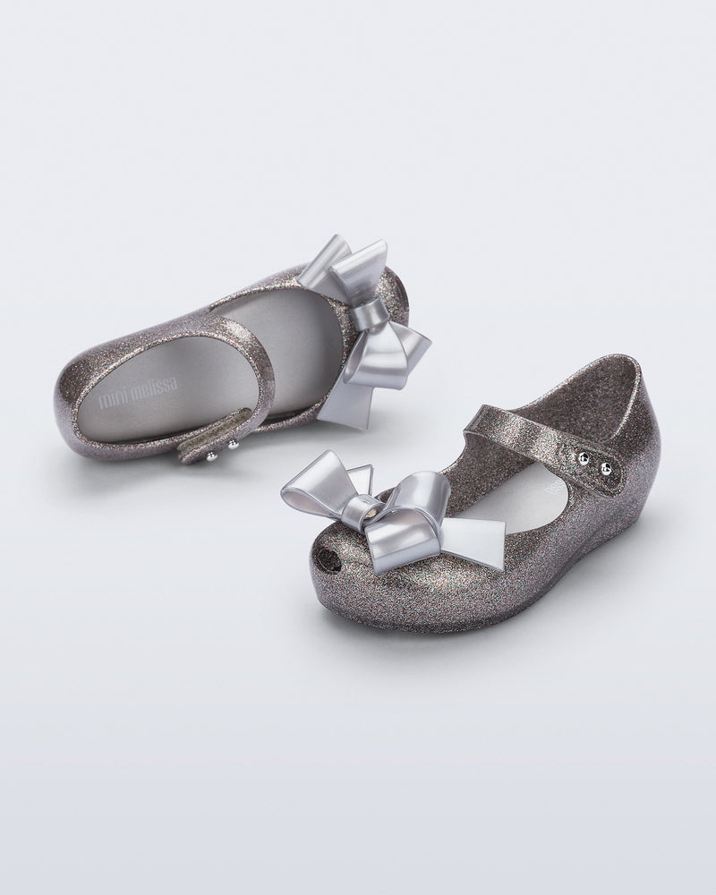 Top and angled view of a pair of silver glitter Ultragirl Classy baby flats with silver bow