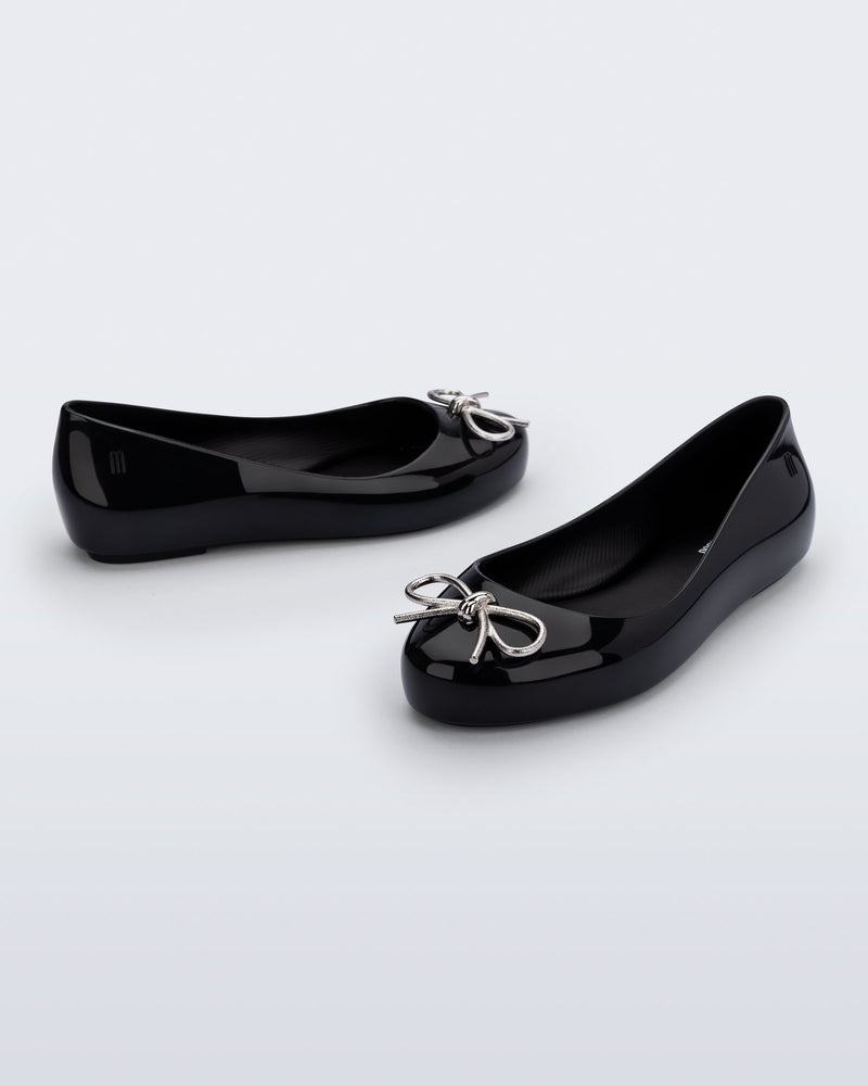 Side and angled view of a pair of black Sweet Love Chrome kids flats with metallic silver bow