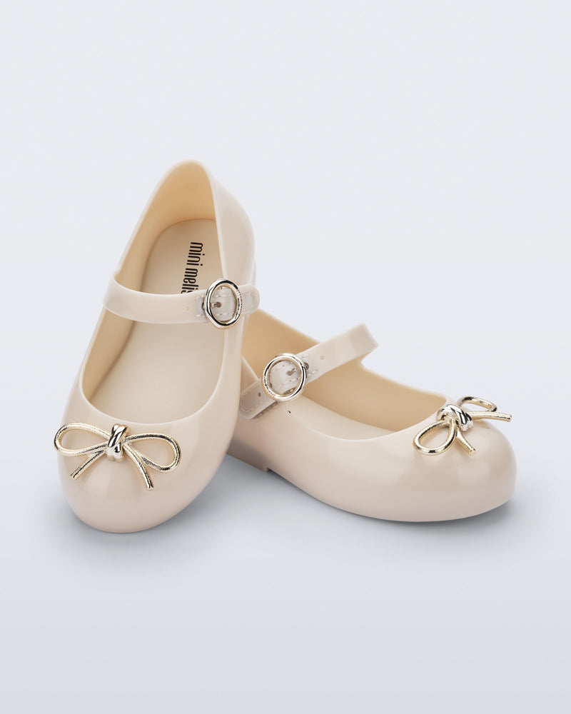Angled view of a pair of beige Sweet Love Chrome baby flats with metallic gold bow and buckle