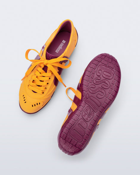 Top and bottom view of a pair of orange-yellow Motion sneakers with pink colored sole and M monogram on side