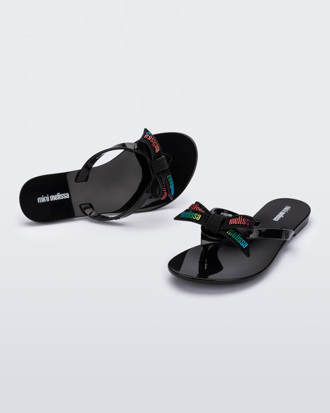 Top and angled view of a pair black Harmonic M Lover kids flip flops with rainbow Melissa logo on bow