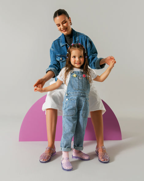 Woman model sitting on a pink shape in a jean jacket and white capris wearing a pair of clear Possession fisherman sandals. Model is holding the hands of a child model standing in front of her wearing overalls and a pair of clear Possession baby fisherman sandals
