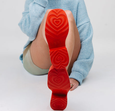 Model in a blue sweater and beige shorts shitting down showing the heart patterned soles of a pair of red Farrah platform mary janes