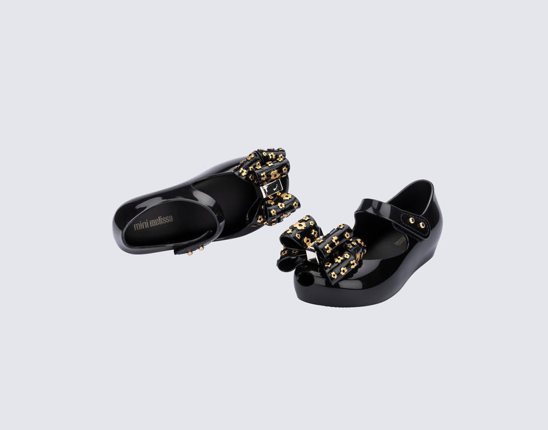 Top and angled view of a pair of black Ultragirl Sweet X baby flats with a bow that has chrome flowers on it