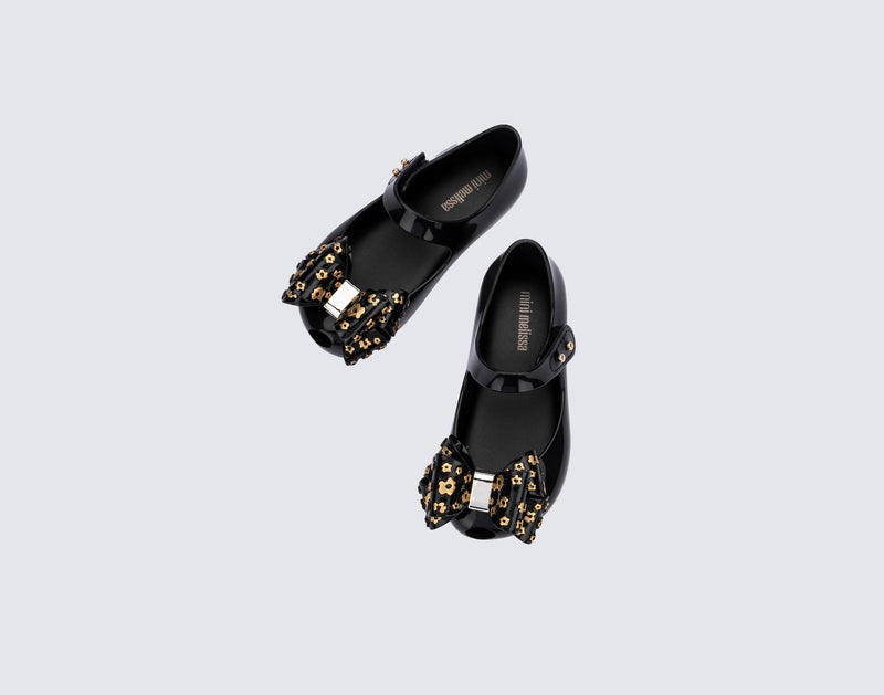 Top view of a pair of black Ultragirl Sweet X baby flats with a bow that has chrome flowers on it