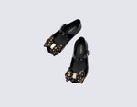 Top view of a pair of black Ultragirl Sweet X baby flats with a bow that has chrome flowers on it