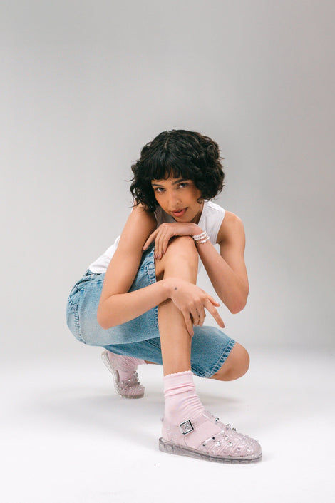 Model in jean shorts and pink socks and a white tank top wearing a pair of clear Possession Stud fisherman sandals