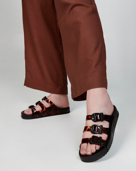 Model's legs in brown pants wearing a pair of black Cozy Stripes slides with tortoise colored straps