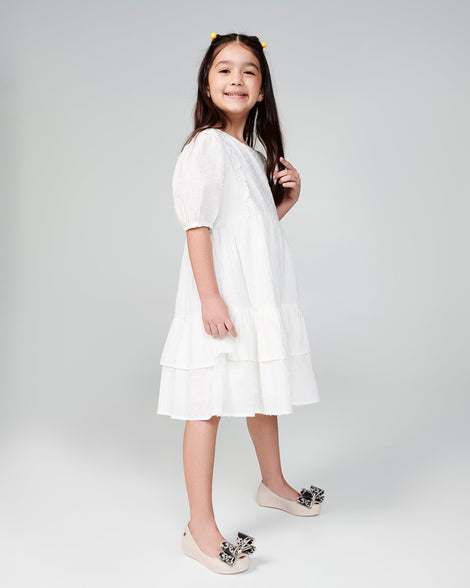 Child model in a white dress wearing a pair of beige kids Ultragirl Classic Bow kids flats with black and clear bow