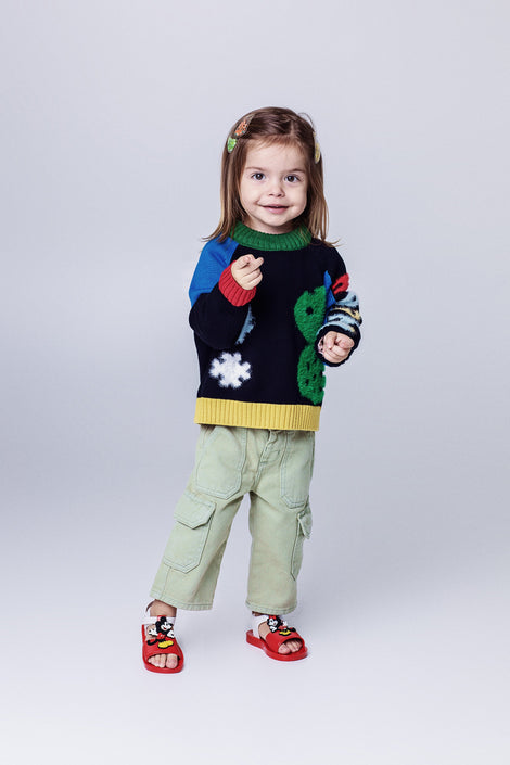 Child model in a sweater and green pants wearing a pair of red Play Mickey & Minnie sandals with white straps