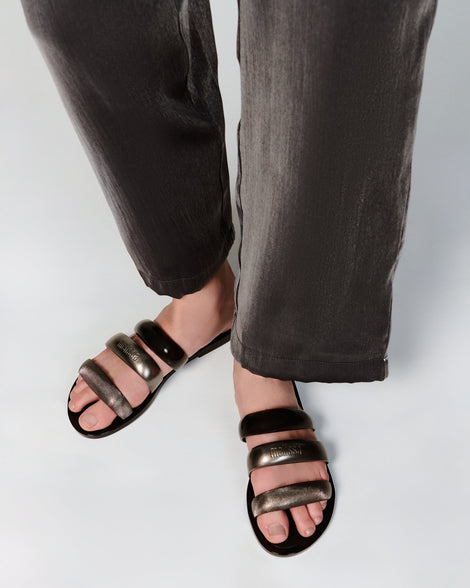 Model's legs in black pants wearing a pair of black Eclipse Slides with gold and black straps.