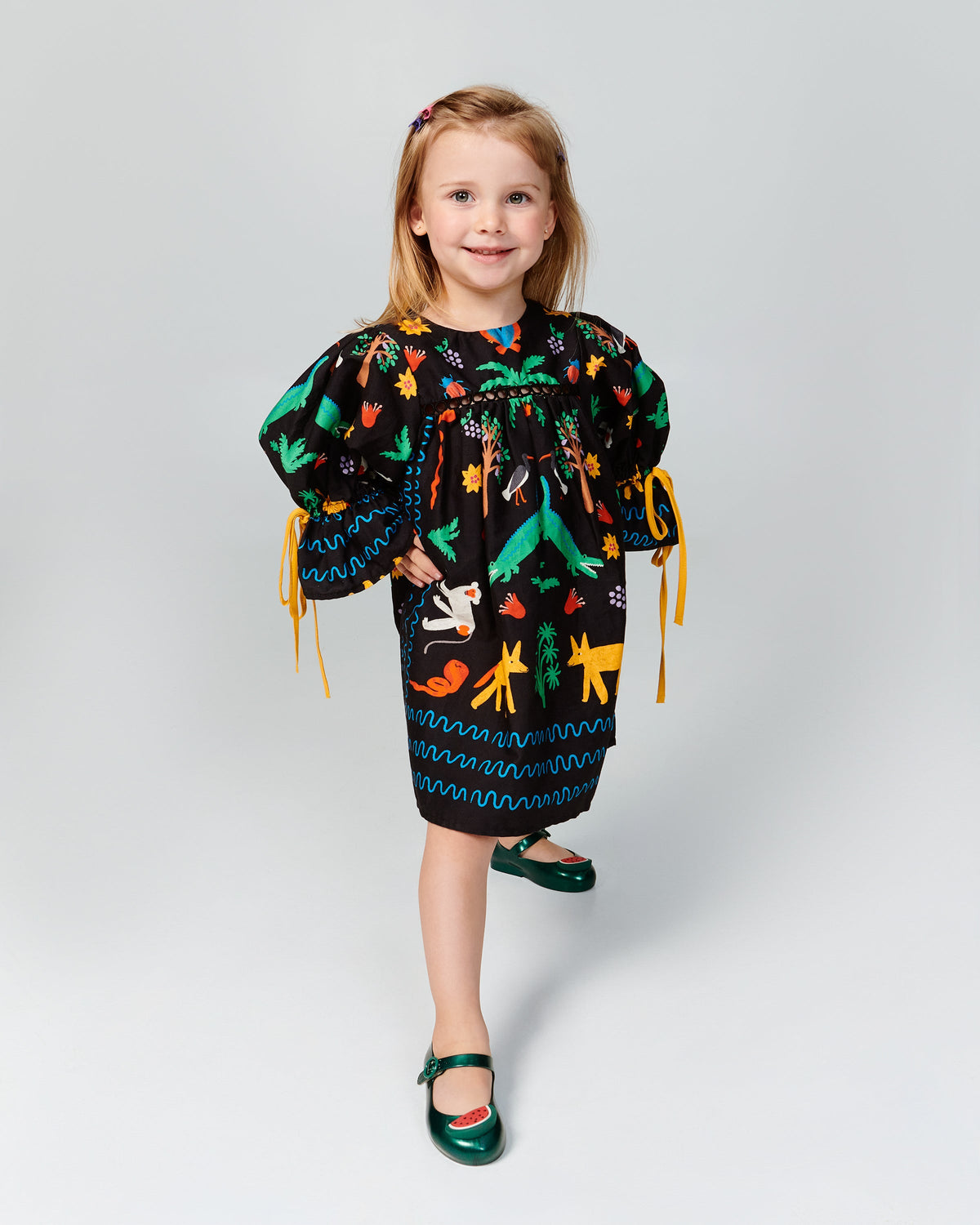 Child model in a black patterned dress wearing a pair of green Sweet Love Picnic baby flats with watermelon slices