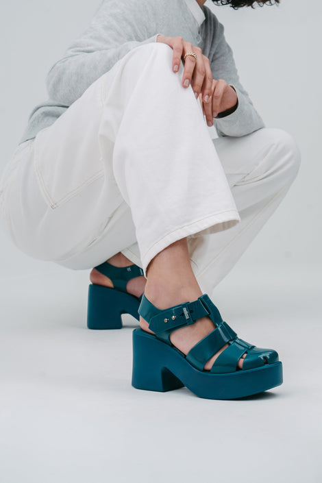A model crouched down wearing a grey sweater and white pants wearing a pair of blue Lulu Platform sandals