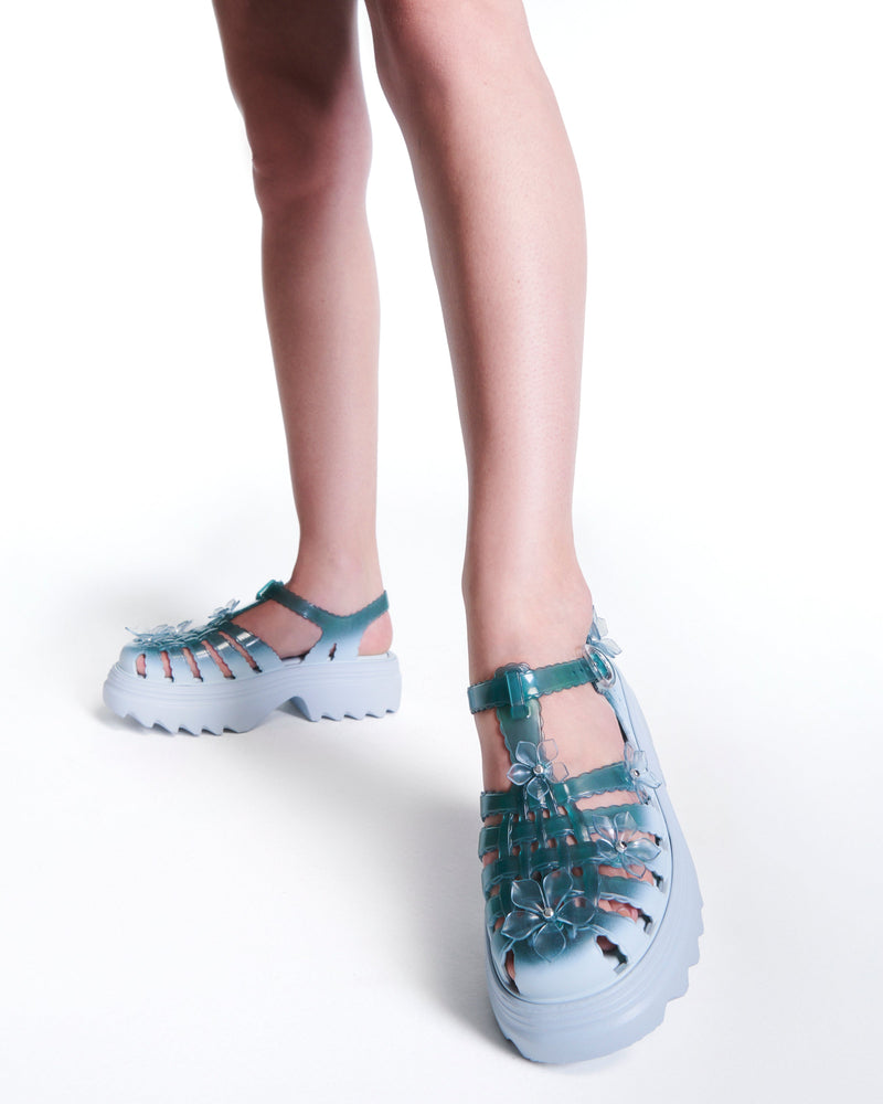 Model's legs wearing a pair of Clear misty blue/green Melissa + Susan Fang Possession Platform Sakura sandals with light blue soles