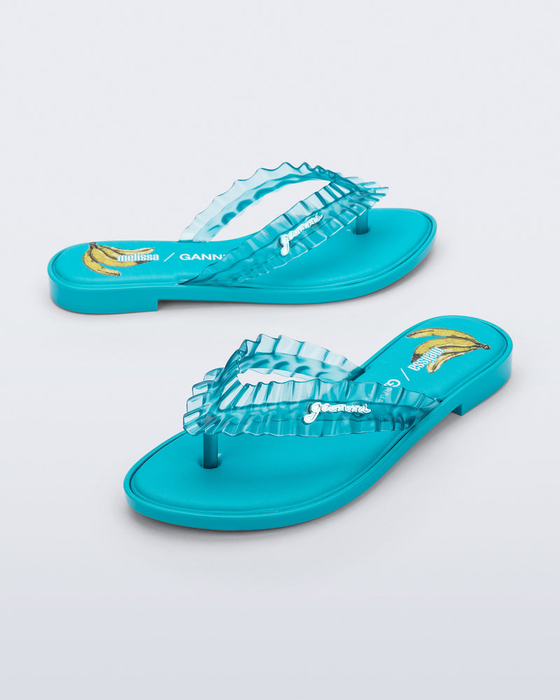 Side and angled view of a pair of blue Flip Flop Slim + GANNI sandals with a banana print foot bed and faux ruffle strap