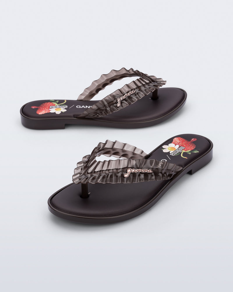 Side and angled view of a pair of brown Flip Flop Slim + GANNI sandals with a strawberry print foot bed and faux ruffle strap