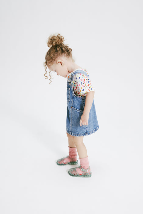 Child model in a jean overall dress wearing star glitter green Possession baby sandals with orange socks