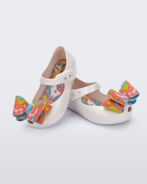 Angled view of a pair of white with multi-colored patterned bow Ultragirl Wonderland baby flats