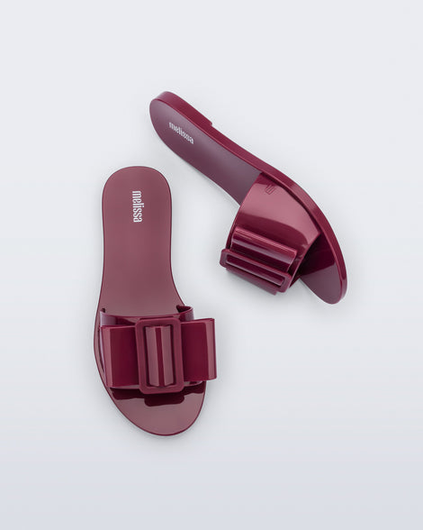 Top and side view a pair of burgundy Babe slide sandals with large bow on top.