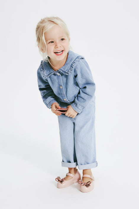 Model in jean one piece holding a pair of sunglasses wearing a pair of pink Sweet Love Glam baby ballerinas with bow