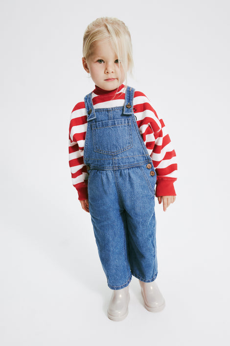 Child model in a red and white strip shirt and jean overalls wearing a pair of pearly beige Chelsea II baby boots