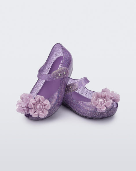 Angled view of a pair clear purple glitter Ultragirl Springtime baby flat with two purple flowers and peep toe.