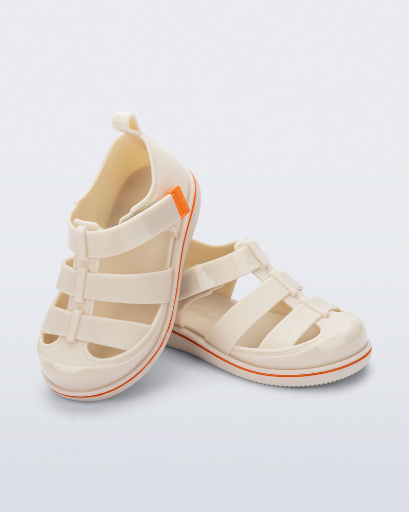 Angled view of a pair of beige Smash closed toe baby sandals with orange trim
