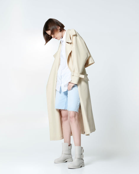 Model in light jean shorts and a white button down with beige rain coat wearing a pair of beige Bang Boots with two side buckles