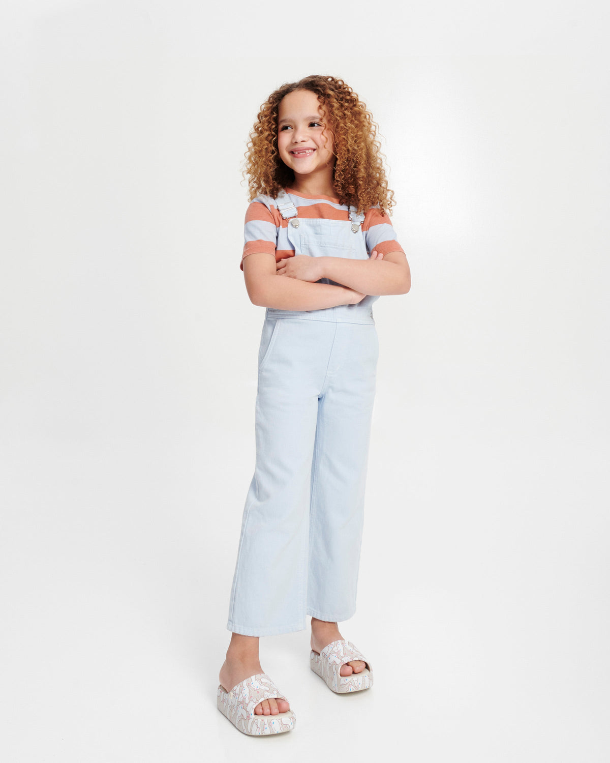 Child model in blue overalls wearing a pair of beige Free Platform slides + Hello Kitty and Friends kids sandals with Hello Kitty and Friends print