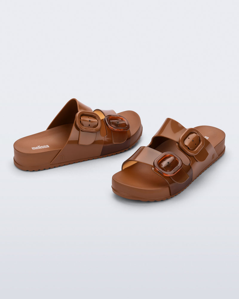 Side and angled view of a pair of brown Cozy slides with part clear brown upper with 2 buckles