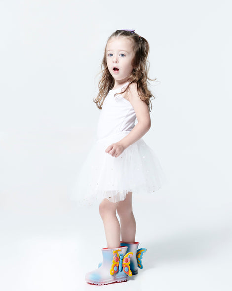 Child model in a white dress wearing a pair of pink with blue splotches Welly Wonderland baby boot with yellow/blue butterfly on side