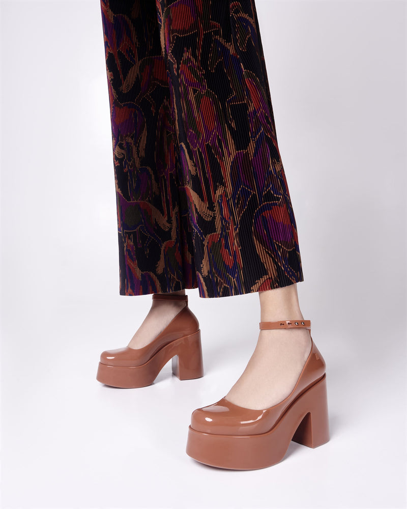 Model's legs in black patterned pants wearing a pair of brown Melissa Doll Heel platforms with ankle strap.