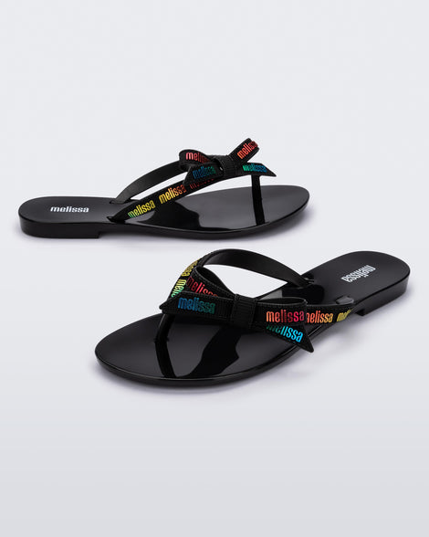 Angled view of a pair of black Harmonic M Lover adult flip flop with rainbow Melissa logo on strap