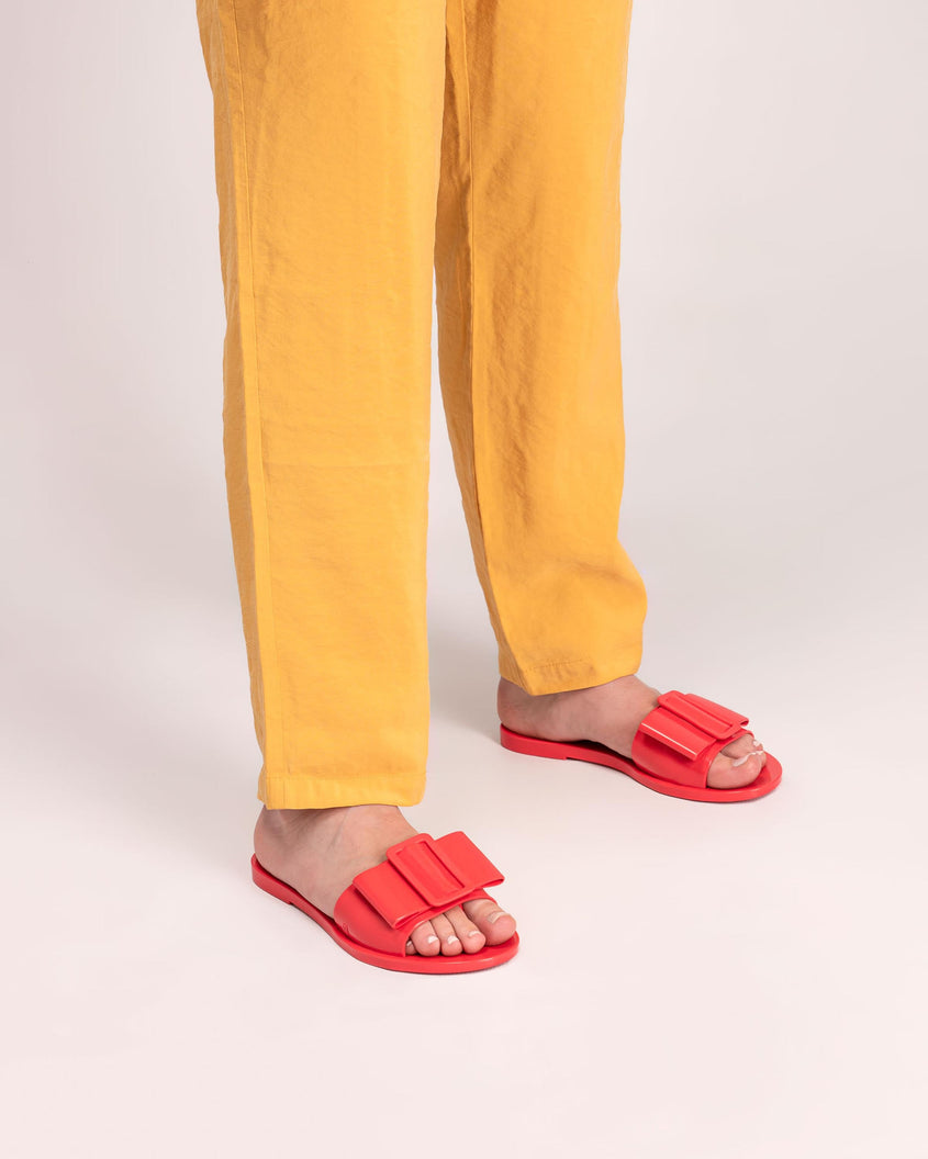 A model's legs wearing yellow pants and a pair of red Melissa Babe slides with a buckle like bow detail on the front strap.