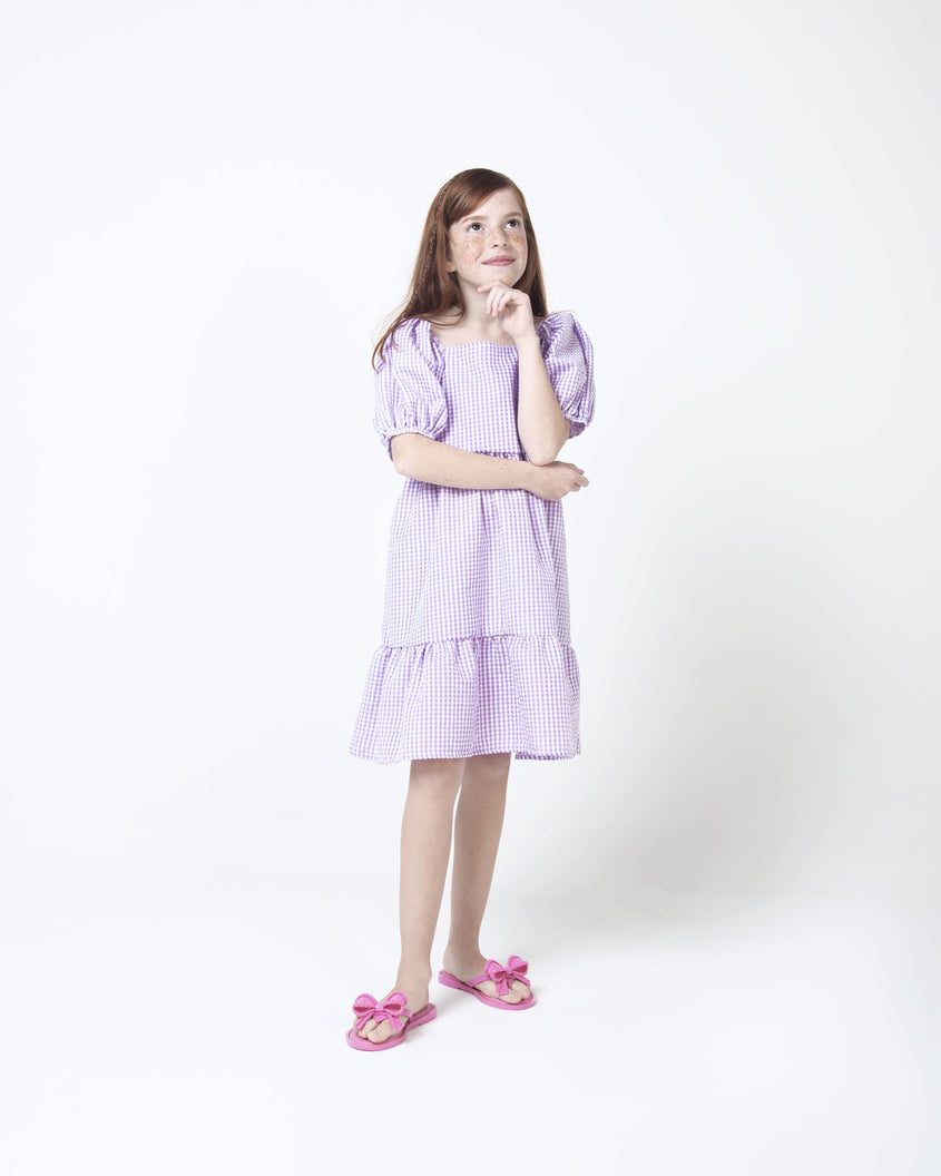A model in a purple gingham dress, posing for a picture and wearing a pair of pink Mini Melissa Slim flip flops with a lace like bow detail on the front strap.