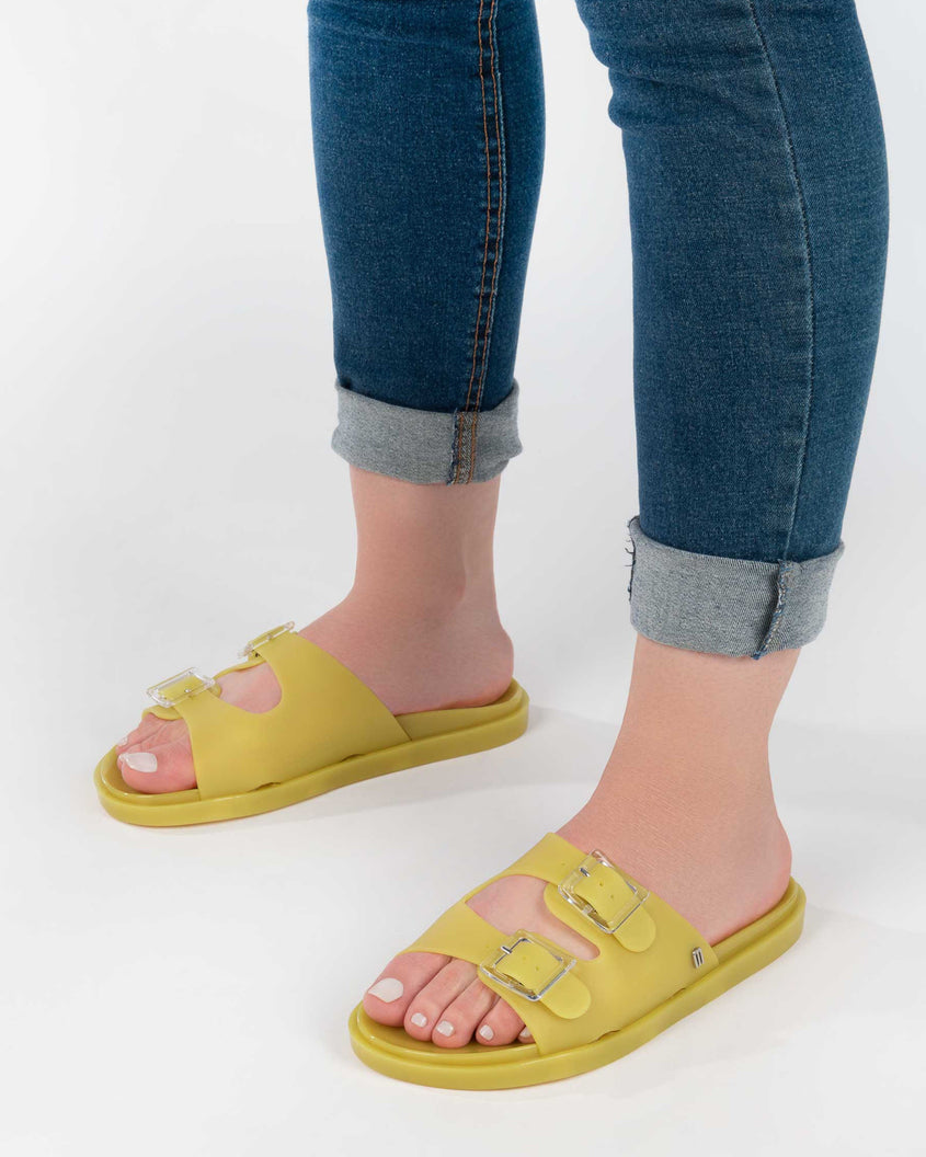 A model's legs wearing jeans and a pair of yellow Melissa Wide Slides with two straps fastened on top with two clear buckles and a