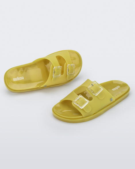 An angled front and top view of a pair of yellow Melissa Wide Slides with two straps fastened on top with two clear buckles and a
