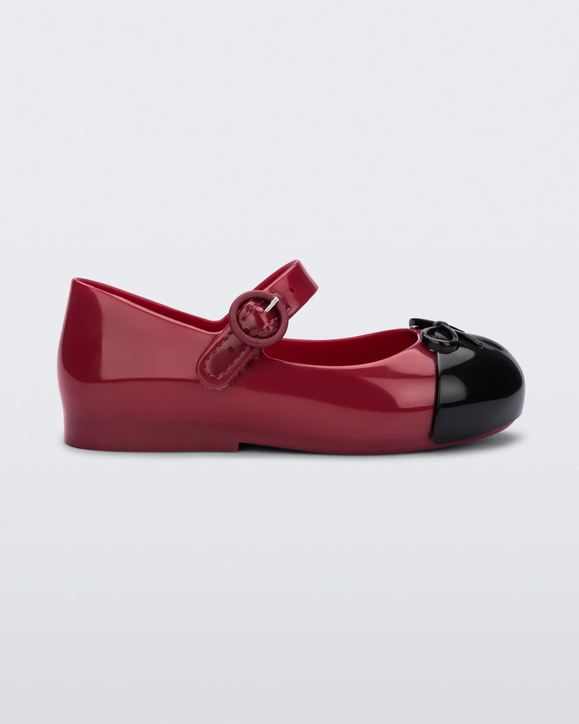 Side view of Red/Black Mini Melissa Sweet Love Cap Toe flat with a red base, top strap and a black toe cap with a bow detail on top.
