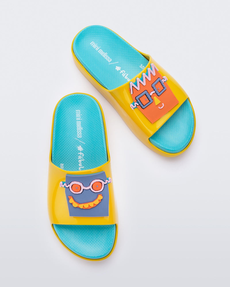 Top view of a pair of yellow/blue Mini Melissa Cloud slides with a blue insole, yellow base and a cartoon drawn smiley face on the front.