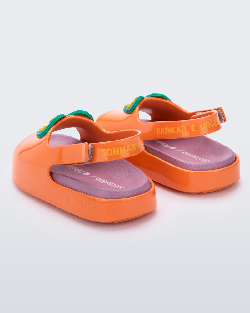 An angled back view of a pair of orange/pink Mini Melissa Cloud sandals with a velcro back strap, pink insole, orange base and a blue/yellow heart drawn on the front.