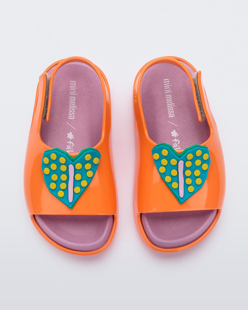 Top view of a pair of orange/pink Mini Melissa Cloud sandals with a velcro back strap, pink insole, orange base and a blue/yellow heart drawn on the front.