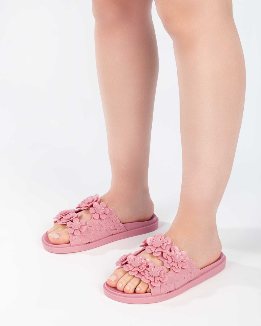 Close up of a model's legs wearing a pair of Melissa Wide Blossom Slides in pink with double floral embossed buckle straps and 3D flower appliques
