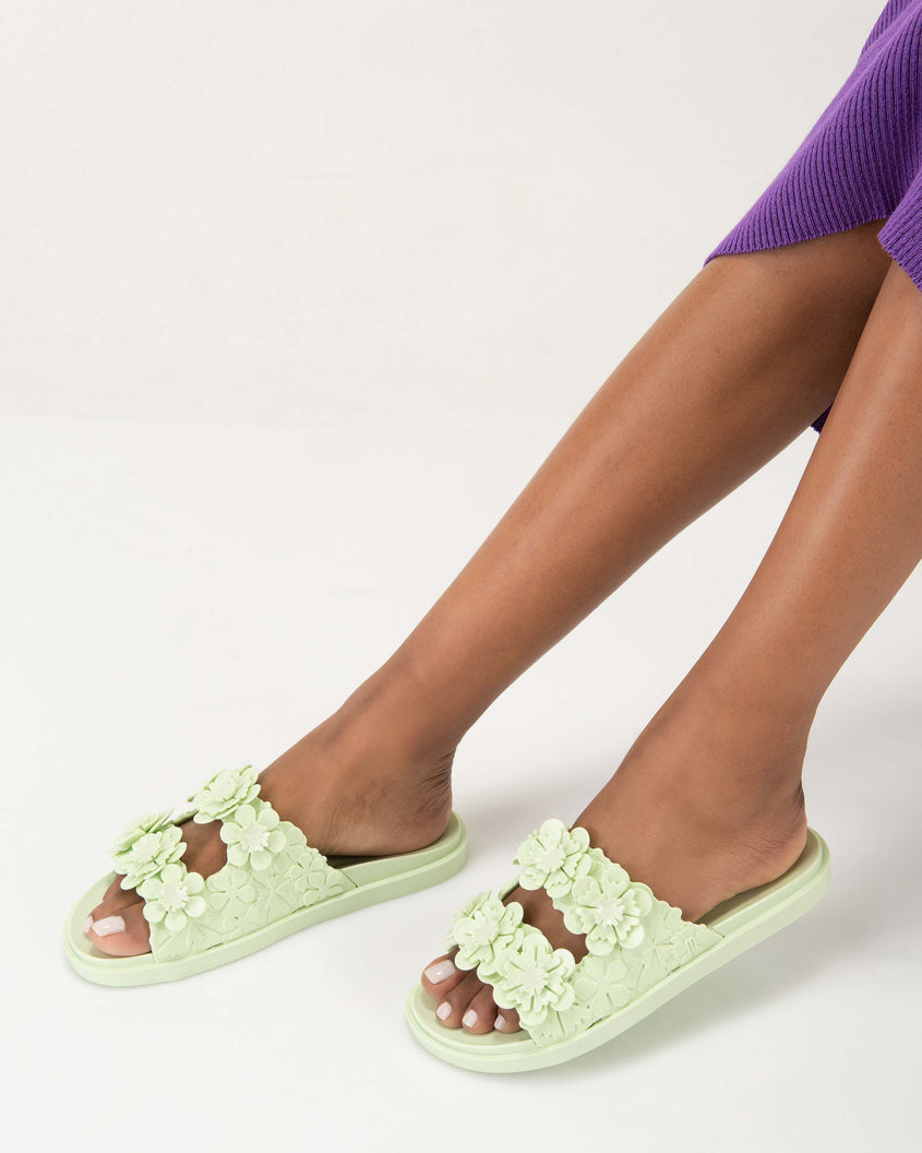 Model's legs wearing a pair of Melissa Wide Blossom Slides in green with double floral embossed buckle straps and 3D flower appliques