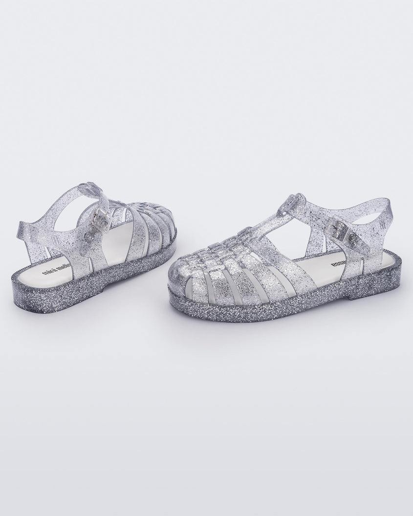 A side and back view of a pair of Glass/Glitter/Sliver Mini Melissa Possession Shiny sandals with several straps and a clear silver glitter base.