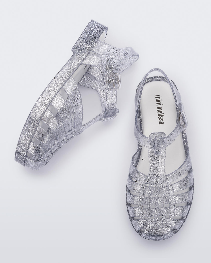 A top and side view of a pair of Glass/Glitter/Sliver Mini Melissa Possession Shiny sandals with several straps and a clear silver glitter base.
