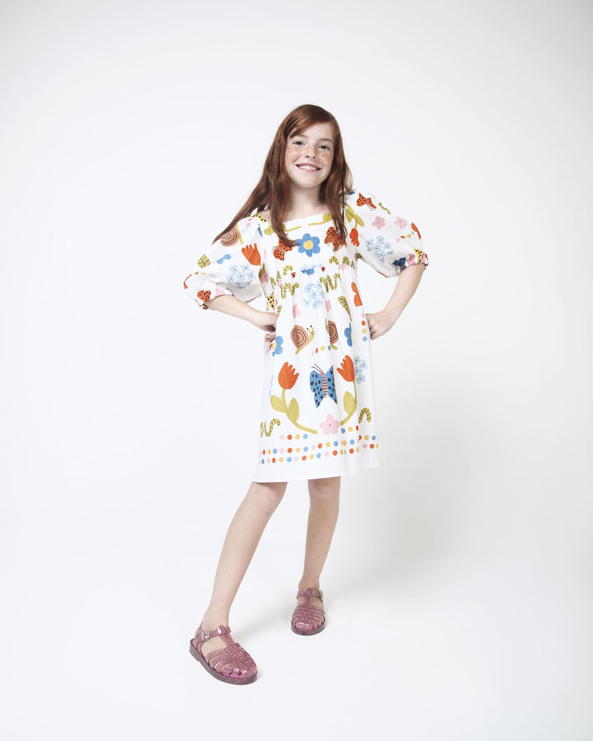 A model in a white patterned dress and a pair Pink/Glitter/Silver Mini Melissa Possession Shiny sandals with several straps and a pink glitter base.