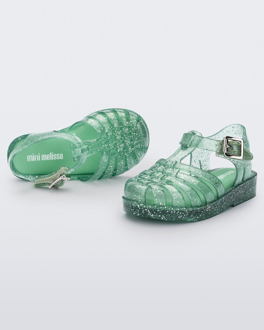 A front and top view of a pair of Green/Glitter/Silver Mini Melissa Possession Shiny sandals with several straps, closed toe front and an ankle strap.