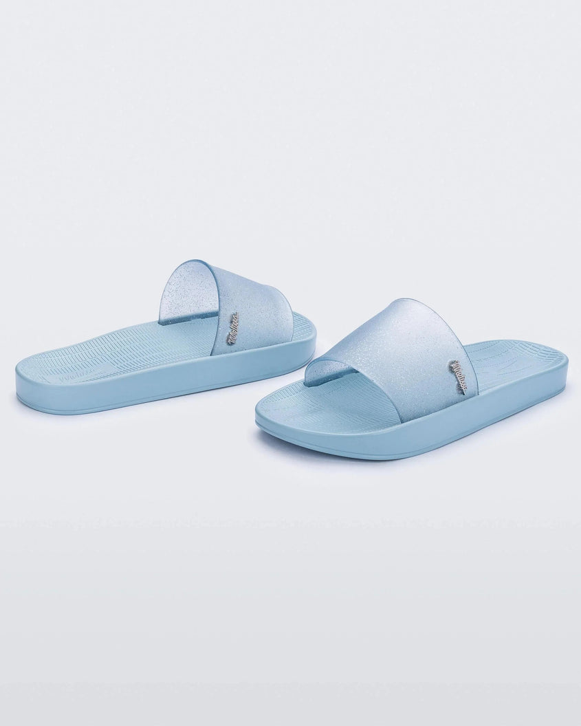 An angled front and back view of a pair of blue/clear glitter Melissa Sun Sunset slides with a blue sole and clear blue glitter front strap.