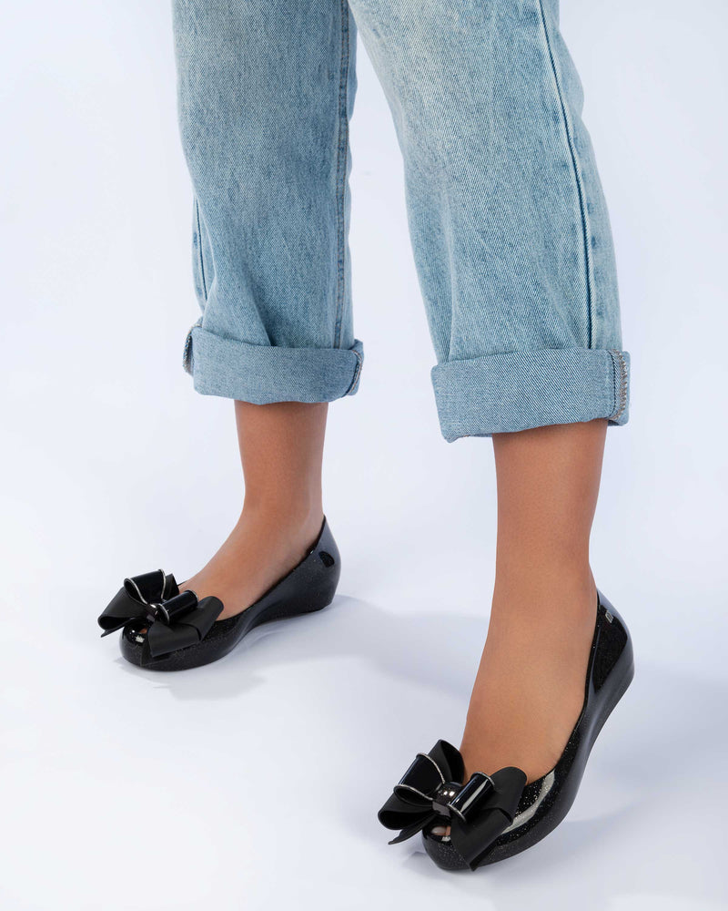 Close up shot of a model's feet wearing the Melissa Ultragirl ballet flats in black glitter colorway. THe model is also wearing a pair of light washed jeans with the hem roled up above the ankle.